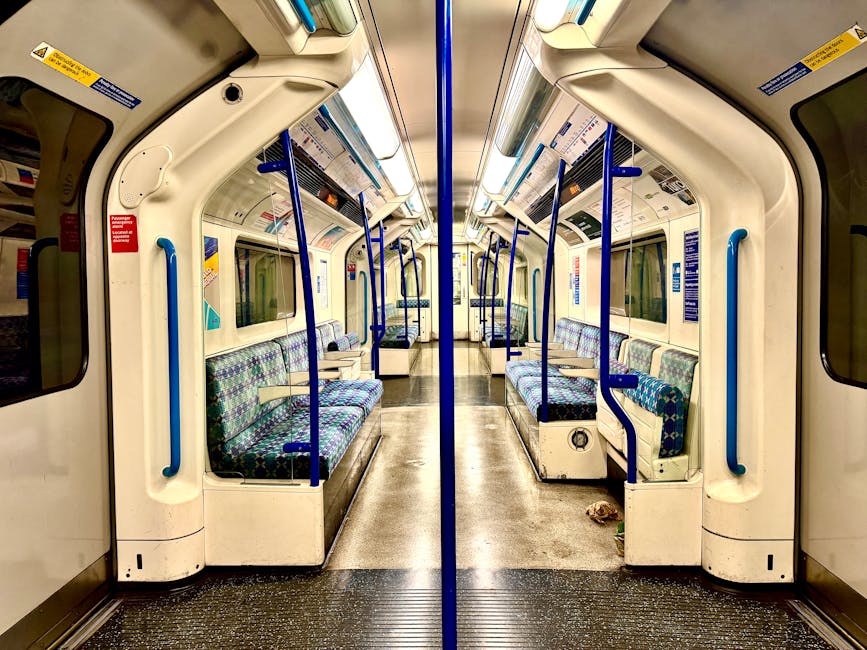An empty interior of a commuter train carriage with beige walls and ceiling, illuminated by natural light through large rectangular windows on both sides. The carriage features blue plastic seats alon